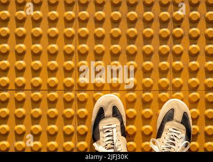 Vista dall'alto delle scarpe da ginnastica su un set di cupole troncate gialle o pavimentazione tattile gialla progettata per aiutare i pedoni con problemi di vista Foto Stock