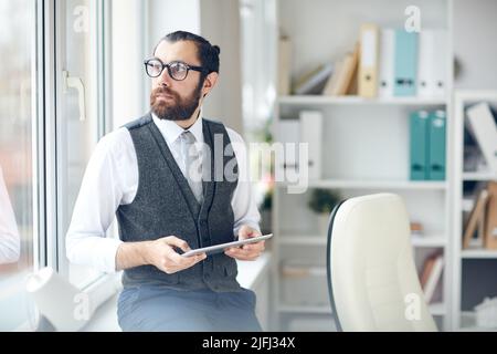 Pensive bel giovane uomo in corpetto formale seduto sul davanzale e guardando fuori finestra mentre si utilizza il tablet e la pianificazione di successo progetto online Foto Stock