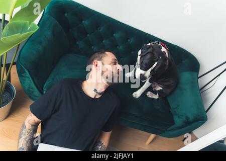 Maschio caucasico freelancer siede sul pavimento con un laptop sul suo grembo mentre amstaff cane lo guarda a casa sfondo medio pieno colpo . Foto di alta qualità Foto Stock