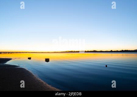 L'alba del porto di Tauranga con sfumature blu e dorate attraverso la tranquilla baia con linee di increspature che si lavano sulla spiaggia. Foto Stock