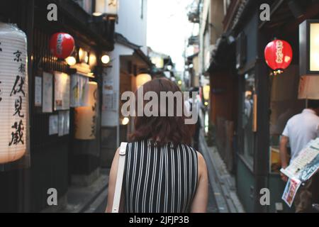 Il retro di una donna giapponese che cammina lungo uno stretto vicolo a Kyoto, Giappone, mentre si guardano negozi e ristoranti. Foto Stock