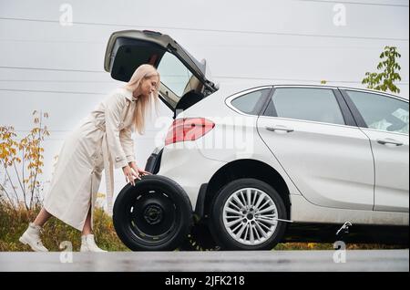 Bella donna che ripara la sua auto bianca con pneumatico forato auto, cercando di cambiare la ruota posteriore. Foto Stock