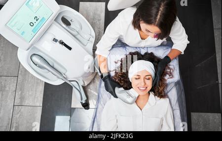 Vista dall'alto della giovane ragazza allegra che si trova sul letto da giorno e sorride mentre riceve il trattamento di cura della pelle facciale. Medico cosmetologo femminile utilizzando un dispositivo a ultrasuoni durante l'esecuzione della procedura di sollevamento del viso. Foto Stock