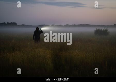 l'uomo in una felpa nera cammina attraverso un prato nella nebbia di notte. Un uomo illumina la strada con una torcia. Foto Stock