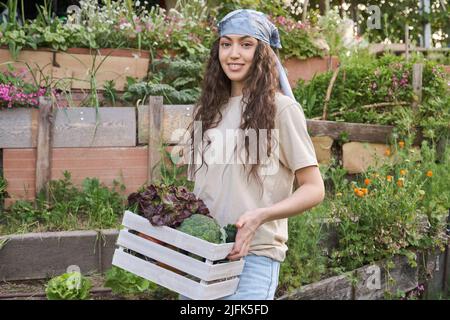 Giovane agricoltore femmina sorridente con un mazzo di verdure in una scatola di legno. Foto Stock