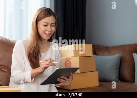 Asian femmina con il proprietario di piccola impresa di riserva che tiene il tablet digitale e le scatole di pacco al dettaglio che controllano l'ordine commerciale di consegna di spedizione sopra Foto Stock