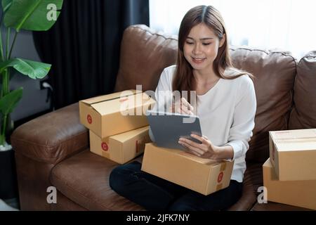 Asian femmina con il proprietario di piccola impresa di riserva che tiene il tablet digitale e le scatole di pacco al dettaglio che controllano l'ordine commerciale di consegna di spedizione sopra Foto Stock