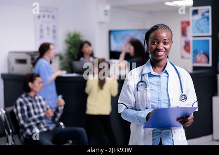 Ritratto di sorridente medico sicuro in occupato ospedale privato in attesa del prossimo appuntamento tenendo appunti. Medic amichevole in camice bianco con stetoscopio in piedi in sala d'attesa clinica. Foto Stock