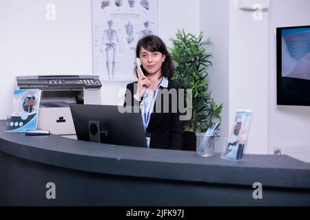 Ritratto del receptionist dell'ospedale sorridente che risponde chiamata dal paziente per prendere un appuntamento per la consultazione clinica in clinica privata. Donna dall'aspetto professionale che lavora alla reception utilizzando il telefono. Foto Stock