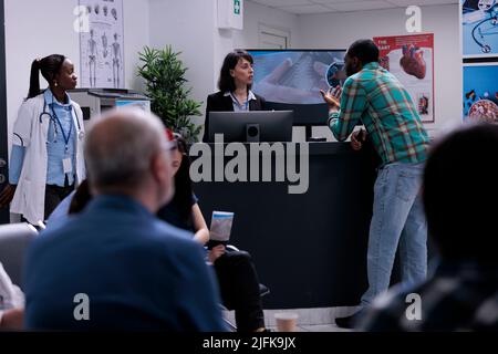 Paziente afroamericano che parla con un receptionist privato della clinica registrandosi per l'appuntamento medico alla reception. Medico in attesa di paziente mentre persone diverse si siedono in sala d'attesa. Foto Stock