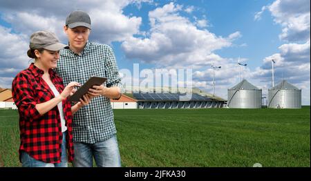 Due agricoltori con computer tablet su uno sfondo di una moderna azienda lattiero-casearia che utilizza energia rinnovabile, pannelli solari e turbine eoliche Foto Stock