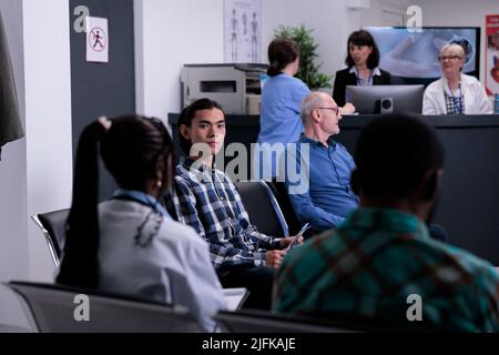 Ritratto di paziente asiatico in attesa di appuntamento medico in ospedale privato, mentre il medico sta parlando con diverse persone. Giovane adulto in clinica medica frequentante medico nomoMet per il controllo. Foto Stock