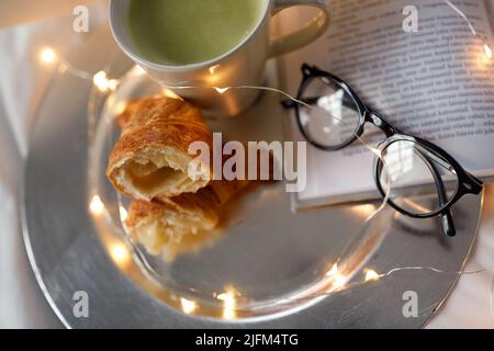 croissant, tè matcha, libro e bicchieri a letto Foto Stock