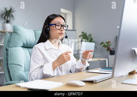 Donna consulente che lavora in remoto utilizzando le cuffie per videochiamate, parlare con i clienti, helpline, donna asiatica che lavora in ufficio. Foto Stock