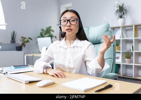 Bella ragazza asiatica di supporto tecnico banca guardando la fotocamera e parlare in videochiamata, utilizzando cuffie, spiegare le informazioni ai clienti, helpline. Foto Stock