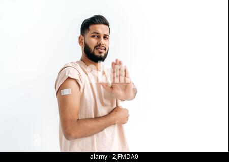 Interrompere la vaccinazione. Il ragazzo indiano o arabo, mostrando cerotto sul braccio dopo la vaccinazione, non voleva essere vaccinato, stare su sfondo bianco isolato, mostra stop gesture con mano Foto Stock