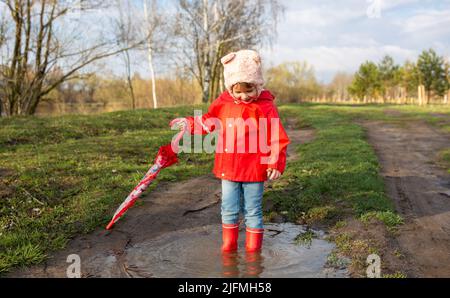 Il bambino gioca con l'ombrello dopo la pioggia in stivali di gomma rossa e un impermeabile. Foto Stock