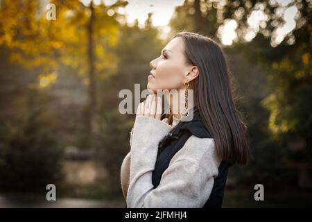 Profilo di una donna seria e pensiva con le mani vicino al viso in autunno all'aperto in giacca beige e giacca senza maniche. Foto Stock