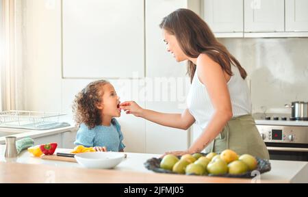 Madre che alimenta le verdure del bambino mentre cucinano insieme nella cucina. Mamma e figlia trascorrono del tempo insieme a casa. Le verdure sono buone per voi Foto Stock