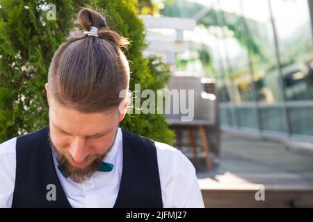 Hairstyle hipster su un giovane uomo barbuto in un vestito seduto in un caffè di strada Foto Stock