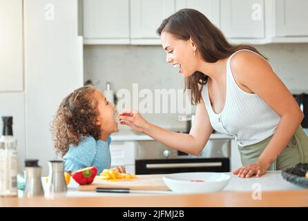 Madre che alimenta le verdure del bambino mentre cucinano insieme nella cucina. Mamma che dice alla figlia di aprire e mangiare le sue verdure Foto Stock