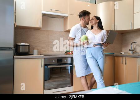 Donna con computer portatile che si appoggia romanticamente al marito in cucina Foto Stock