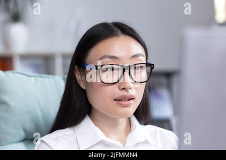 Primo piano ritratto fotografico di una giovane donna asiatica in ufficio, indossando occhiali e una camicia bianca, lavorando in ufficio, guardando il monitor. Foto Stock