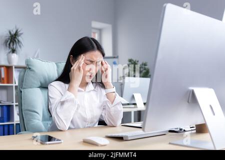 Donna asiatica d'affari ammalata che lavora in ufficio, avendo mal di testa grave, tenendo le mani sulla testa in depressione e triste. Foto Stock