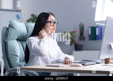 Lavoratore di ufficio donna asiatica stanca, ha dolore al collo severo, massaggia i muscoli del collo con la mano, lavoratrice femminile in ufficio al calcolatore. Foto Stock