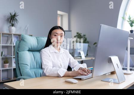 Ritratto di una giovane donna d'affari di successo, un lavoratore in un ufficio moderno che lavora ad un computer, una donna asiatica che guarda la macchina fotografica. Foto Stock