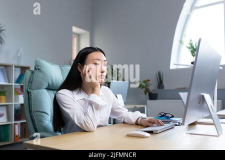 Una triste donna d'affari lavora in un ufficio moderno, una donna asiatica pensa ai risultati del lavoro. Foto Stock