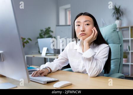 Una triste donna d'affari lavora in un ufficio moderno, una donna asiatica pensa ai risultati del lavoro. Foto Stock