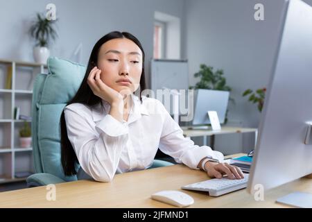 Una triste donna d'affari lavora in un ufficio moderno, una donna asiatica pensa ai risultati del lavoro. Foto Stock