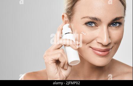 Donna caucasica positiva applica crema idratante sul suo viso, su sfondo grigio. Crema per il viso pelle idratante, cura della pelle Foto Stock