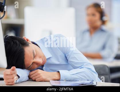 Un agente del call center asiatico esausto che dorme alla sua scrivania in un ufficio. Uomo d'affari che si sente sovraccaricato, stanco e demotivato durante il funzionamento Foto Stock