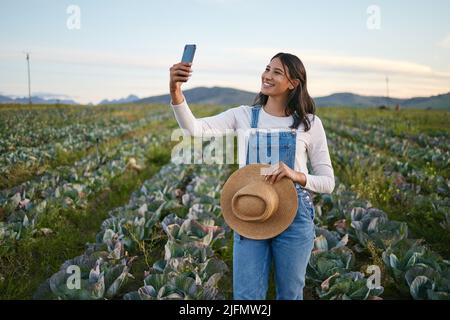 Donna agricoltore che prende un selfie sul suo smartphone mentre si trova in un campo di cavolo. Giovane donna brunetta con un cappello di paglia che utilizza il suo dispositivo mobile su un Foto Stock