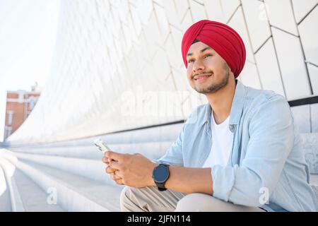 Sereno uomo indiano sorridente in testiera nazionale turban seduto sui gradini all'aperto, tenere smartphone, trascorrere il tempo in social network, messaggistica e chat online, guarda via da sogno Foto Stock