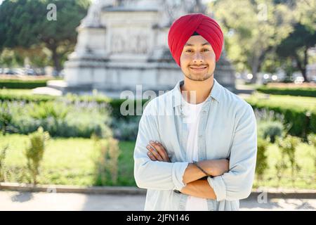 Bel giovane uomo indiano sorridente che indossa il tradizionale turbante rosso si erge all'aperto con le braccia ripiegate. Fiero uomo sikh sicuro con le braccia incrociate guardando la macchina fotografica in piedi in paesaggio urbano Foto Stock