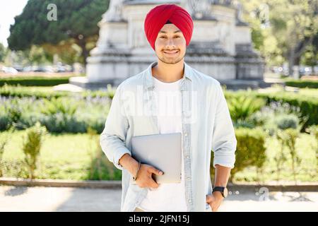 Sorridente uomo indiano positivo in tradizionale headwrap tumban che tiene il laptop in piedi all'aperto, ritratto di maschio studente indù, imprenditore con computer portatile in cityscape Foto Stock