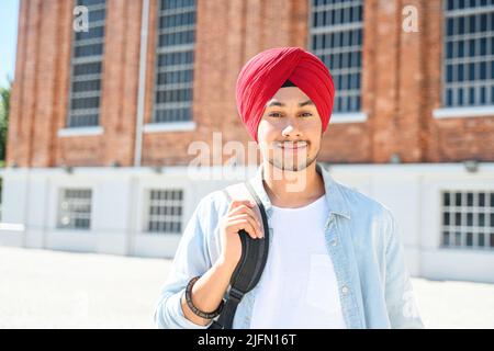 Ritratto di giovane studente indiano giovane felice che indossa tradizionale rosso turban pagg e maglietta casual nel campus, ragazzo hindu ottimista sorridente con zaino in piedi all'aperto vicino all'università Foto Stock
