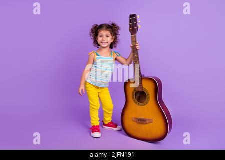 Foto integrale del corpo di ragazza piccola bruna divertente con chitarra indossare pantaloni scarpe top isolato su sfondo viola colore Foto Stock