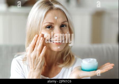 Close-op di positivo caucasica bionda donna cura per la pelle del viso, utilizzando il giorno idratante, mezza età femmina applicazione anti-invecchiamento crema cosmetica per nascondere le rughe, facendo una routine quotidiana di bellezza, sorride Foto Stock