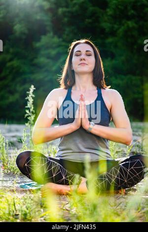 una donna pratica yoga e medita in posizione loto sulla sabbia sullo sfondo di alberi Foto Stock