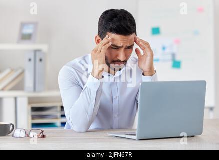 Giovane uomo d'affari misto che soffre di mal di testa lavorando su un notebook in un ufficio solo al lavoro. Un uomo ispanico che si sente stressato mentre lavora Foto Stock