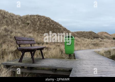 Cestino verde chiaro con pittogramma accanto a una panchina sul lungomare attraverso le dune di sabbia di Amrum (Germania) in un pomeriggio d'inverno Foto Stock