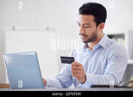 Giovane serio bella gara mista uomo d'affari che tiene e utilizzando una carta di credito e di lavoro su un portatile in un ufficio solo al lavoro. Boss maschio ispanico Foto Stock