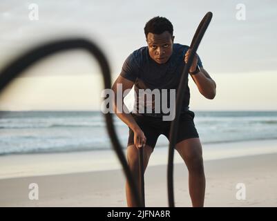 Uomo che fa l'allenamento utilizzando due corde di battaglia sulla spiaggia. Fit uomo afroamericano che esercita sul mare. Dedicato al fitness Foto Stock