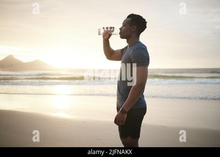 Montare l'uomo che beve acqua, facendo una pausa. Giovane atleta afroamericano che si esercita sulla spiaggia. Uomo serio messo a fuoco che beve da una bottiglia. Sicuro Foto Stock