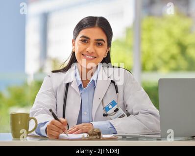 Ritratto di un giovane medico indiano che indossa uno stetoscopio seduto in un ufficio scrivendo una prescrizione mentre si siede alla sua scrivania. Sorridente femmina gp o. Foto Stock