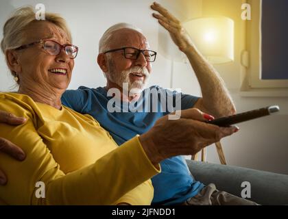 Felice coppia anziana seduta sul divano a casa, guardando la tv e ridendo Foto Stock